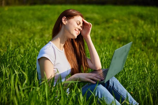 a woman works at a laptop sitting in a field covering her face from the sun with her hand. High quality photo