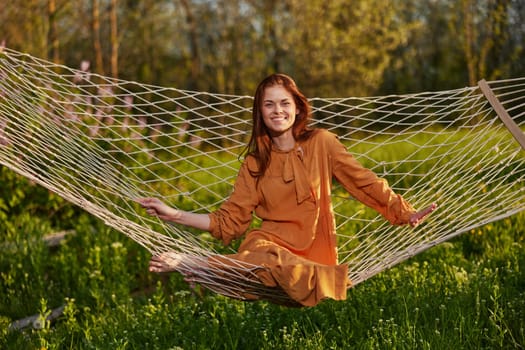 horizontal photo of a beautiful, red-haired woman lying in a hammock enjoying a rest in a long orange dress, on a warm summer day, smiling happily looking at the camera. High quality photo