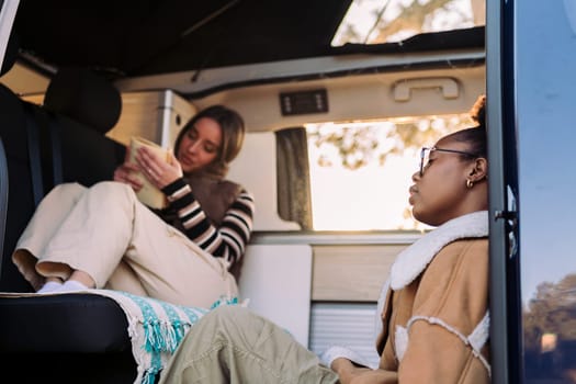 two young friends chilling and reading during a road trip in camper van, concept of travel adventure and female friendship