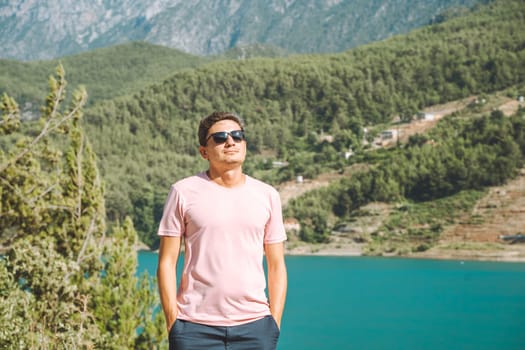 Portrait of Smiling man in sunglasses standing near mountains lake on background. Positive young male traveling on blue lake outdoors travel adventure vacation.