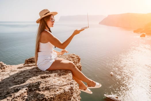 Working remotely on seashore. Happy successful woman female freelancer in straw hat working on laptop by the sea at sunset. Freelance, remote work on vacation