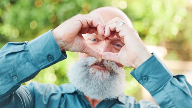 Portrait, hands and heart with a senior man outdoor in a garden during summer for love or health. Face, emoji and shape with a hand gesture by a mature male outside in a park for wellness or romance.