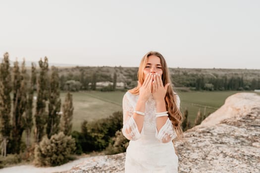 Romantic beautiful bride in white dress posing with sea and mountains in background. Stylish bride standing back on beautiful landscape of sea and mountains on sunset