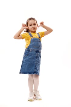 Full length vertical portrait of lovely little girl wearing bright yellow t-shirt and denim sundress, smiling cutely and showing thumbs up, looking at camera, isolated over white background