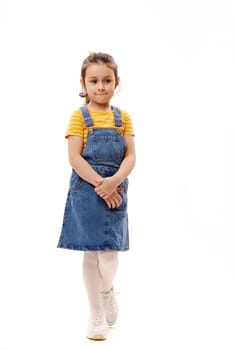 Full length vertical portrait of Caucasian lovely little preschooler child girl in yellow t-shirt and blue denim sundress, smiling cutely, looking at camera, isolated over white background.