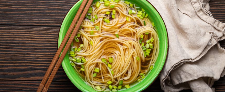 Asian noodles soup in green rustic ceramic bowl with wooden chopsticks top view on dark wooden background. Lo mein noodles with broth and green onion