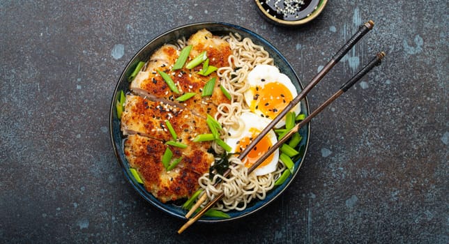 Asian noodles ramen soup with deep fried panko chicken fillet and boiled eggs in ceramic bowl with chop sticks and soy sauce on stone rustic background top view