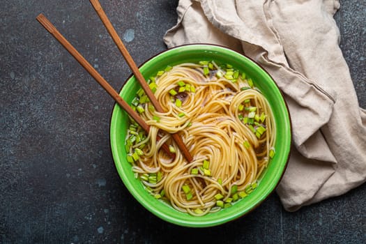 Asian noodles soup in green rustic ceramic bowl with wooden chopsticks top view on rustic stone background. Lo mein noodles with bouillon and green onion