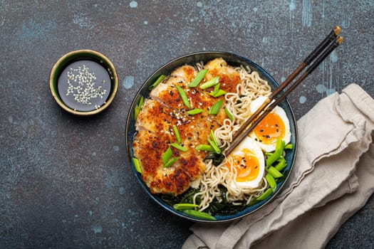 Asian noodles ramen soup with deep fried panko chicken fillet and boiled eggs in ceramic bowl with chop sticks and soy sauce on stone rustic background top view