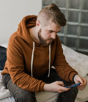 Freelance man using mobile phone checking social media network feed or message chat sitting on bed at home. Online digital communication rest after hard work day