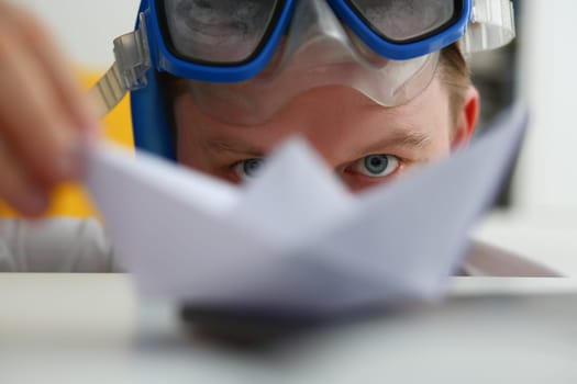 Young man in swimming goggles plays with paper ship at workplace. Tourism resort idea cruise ship ticket sales concept