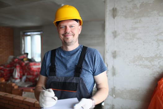 Joyful handyman, builder in uniform and hard hat looks at camera and takes notes on clipboard. Builder foreman preparing for repair work