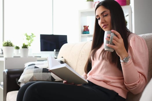 Woman is reading book and drinking from cup sitting on couch. Home leisure and reading books concept