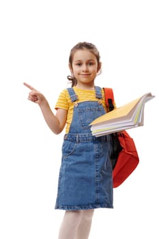 Positive smart schoolgirl, first grader holding textbooks, pointing at copy ad space on isolated white background, smiling looking at camera. Welcome to new semester of academic year. Back to school