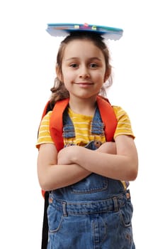 Authentic portrait Caucasian 5-6 years old positive cute child girl, first grader with book on her head, smiling cutely, looking at camera, posing with her arms crossed over isolated white background