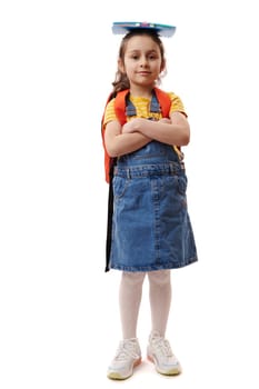 Full size portrait of lovely smart school child girl, first grader with book on her head, smiling cutely, looking at camera, posing with her arms crossed over isolated white background. Back to school