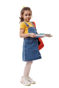 Full length portrait of Caucasian smiling excellent best student, first grader schoolgirl, 5-6 years old little girl holding books and copybooks going to school, isolated on white background. Ad space