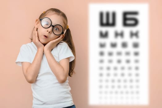 Surprised girl 5 - 7 years old in round glasses against the background of a blurred sivtsev table for checking visual acuity.