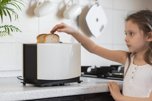 Kid girl cooking on kitchen. White toaster with sliced bread stands on the work surface of the kitchen table.