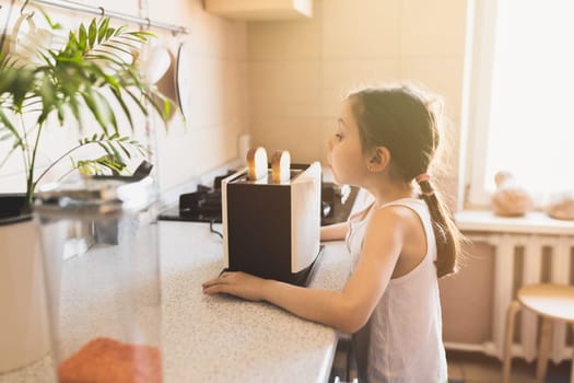 Funny caucasian girl 6 years old in a home kitchen looks with surprise at a toaster with bread on a working kitchen tabl, selective soft focus.