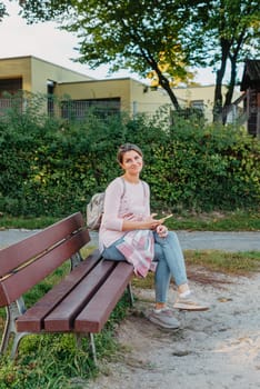 Girl sitting on a bench and watching distant city scenic. Girl enjoying city view from a bench in sunset or sunrise time. Beautiful blond-haired pretty girl in summer green park against sunset beams. Lonely woman sitting alone moments sunset. On Blurred background bokeh. light Fair.