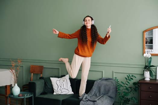 A happy, pretty young woman wearing headphones dances to her favorite song at home. Cheerful funny girl listening to popular music, moving, jumping on the couch in the living room, experiencing joy