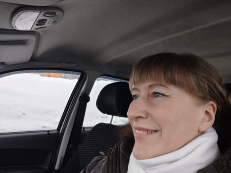 Portrait of adult cute funny blonde woman in warm clothes taking a selfie inside the car and driving it. The concept of pleasant trip in spring, autumn, winter