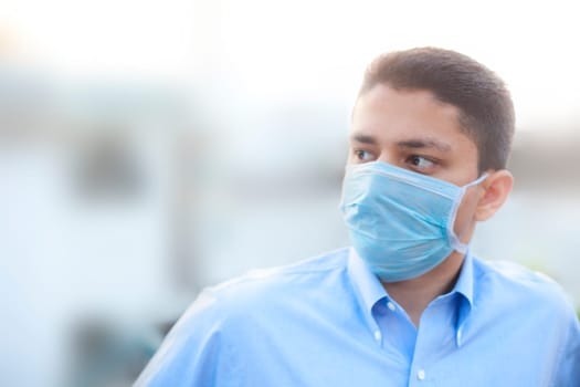 Portrait shot of a young man in a blue colored shirt and wearing a surgical mask or a procedure mask with blurred background.