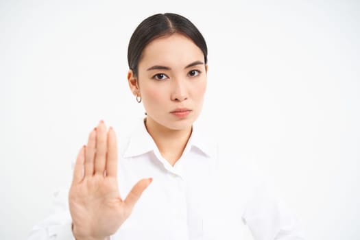 Please stop, enough. Serious asian female manager, businesswoman shows extended hand gesture, disapproves, prohibits smth, white background.