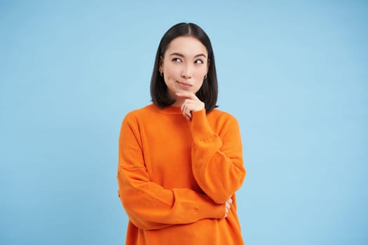 Brainstorm, ideas concept. Portrait of asian girl thinks and looks puzzled, stands thoughtful in orange sweatshirt, makes decision, blue background.
