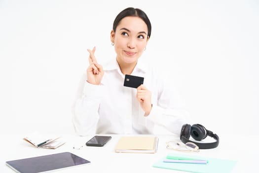 Hopeful asian woman, office worker shows credit card and cross fingers for good luck, makes wish, white background.