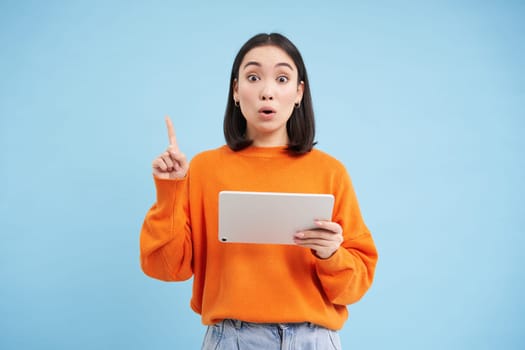 Eureka, got an idea. Excited asian woman with digital tablet, raising finger up, got revelation, has great solution, stands over blue background.