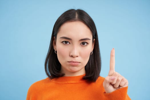 Close up of serious asian woman shows one finger, stop gesture, scolding, disapprove smth, stands over blue background.