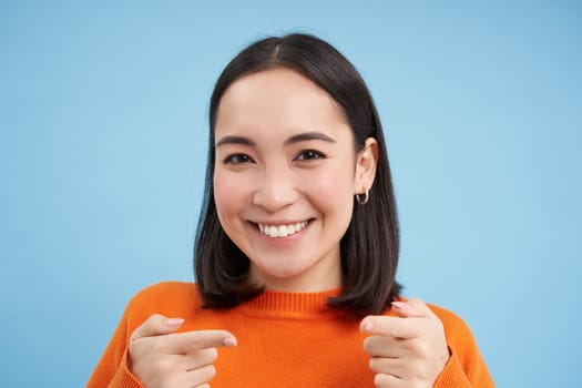 Gotcha. Close up portrait of smiling asian woman points fingers at camera pleased, congratulates you, stands over blue background.