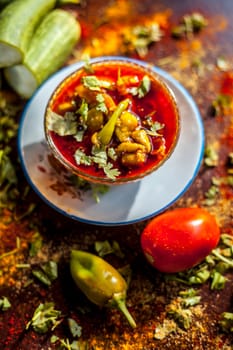 Close-up shot of fresh Indian Lunchtime dish luffa or Galka nu Shaak in a glass container along with all its ingredients on a rust-colored surface.