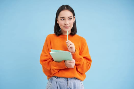 Thoughtful asian woman, student smiling, holding pen and notebook, thinking, inspired to write something, standing over blue background.