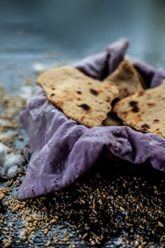 Close-up shot of round bread Bhakri on the black wooden surface along with some raw whole wheat, and salt in a container. Shot of Bhakri in a container on the black surface.