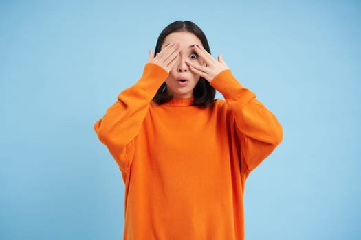 Portrait of cheerful cute asian girl waits for surprise, shuts her eyes with hands, birthday girl awaiting for gift, stands over blue background.