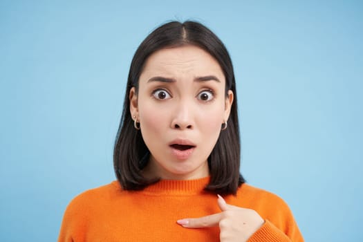 Close up of shocked asian girl, points at herself with offended look, stands insulted over blue background.