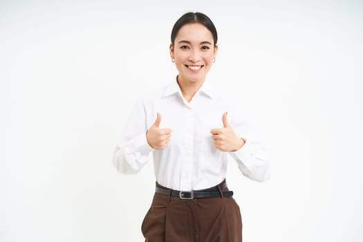 Smiling asian woman shows thumbs up, approves promo offer, recommends product, stands isolated over white background.