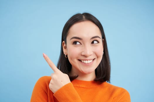 Close up shot of smiling asian girl, points finger at upper left corner, shows banner, demonstrates advertisement, blue background.