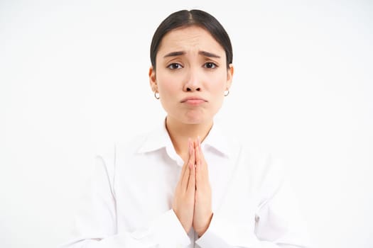 Upset young saleswoman, asian woman holds hands in pray, begs you, asks for favour with sad face, pleading, standing over white background.