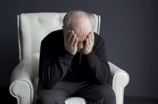 A man in a black turtleneck is sitting in a white chair, rubbing his face with his hands, holding his head. Depression, headache. Medical concept.