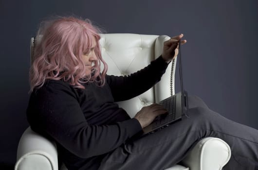 A man in a pink wig sits in a white chair and communicates via video link on a laptop. Side view