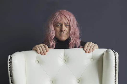 Portrait of a man in a pink wig, who is leaning on a white chair, looking at the camera, posing. close-up.