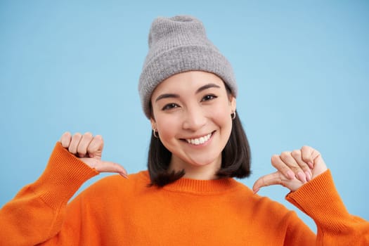 Close up portrait of confident korean woman in warm hat, points at herself with self-assured face, standing over blue background.