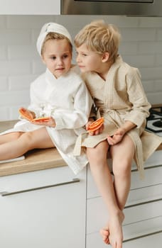 A boy and a girl in bathrobes are sitting in the kitchen talking, eating candied fruit.