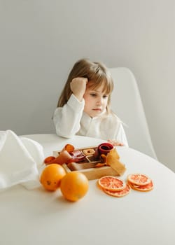 A girl in a white coat sits at a table, looks away, is going to eat marmalade.