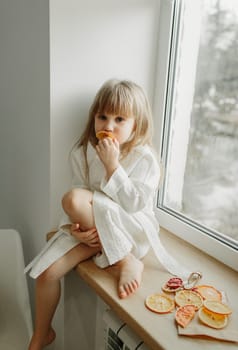 A girl in a white coat is sad sitting on the windowsill, eating fruit marshmallow.
