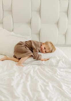 A boy in a brown bathrobe, lies on a white bed, covers his face with his hands.